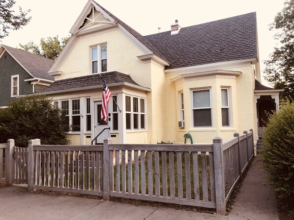 Une maison à deux étages avec un toit à pignon, un porche à l’avant et une clôture en piquets.