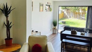 A modern living room with a sofa, a coffee table, and a dining table. There is a large window with a view of a backyard. - 3 By The Sea, Ground Floor Apartment By The Sea (Worthing)