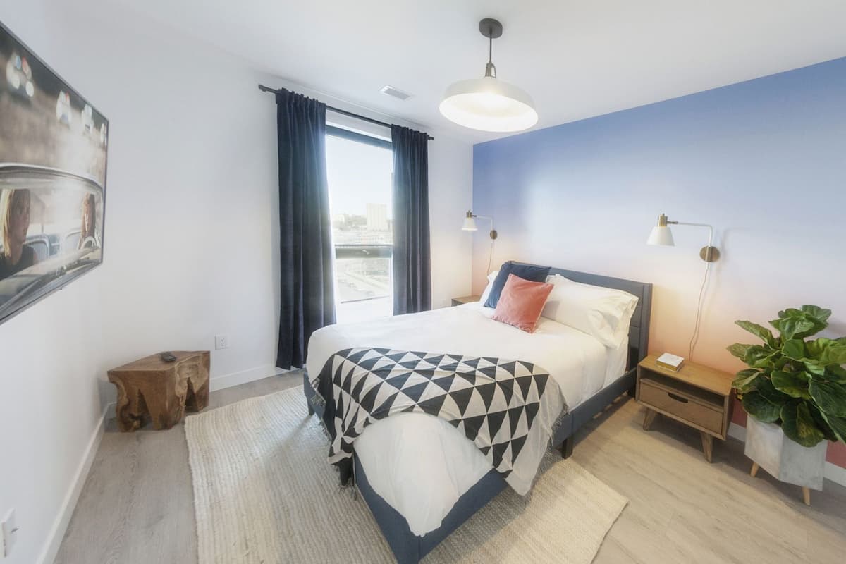 A modern bedroom with a bed, bedside tables, a nightstand, a hanging light, a plant, and a window with curtains.