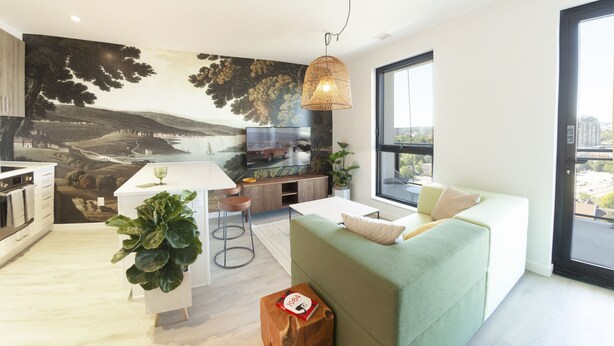 Ein modernes Wohnzimmer mit einem grünen Sofa, einem Esstisch und einem großen Fernseher. An der Wand hängt ein Gemälde und in der Ecke steht eine Pflanze.