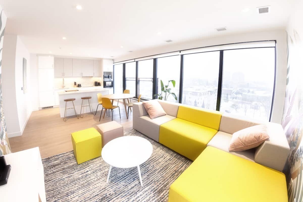 A modern living room with a large window, a kitchen in the background, and a yellow sofa.