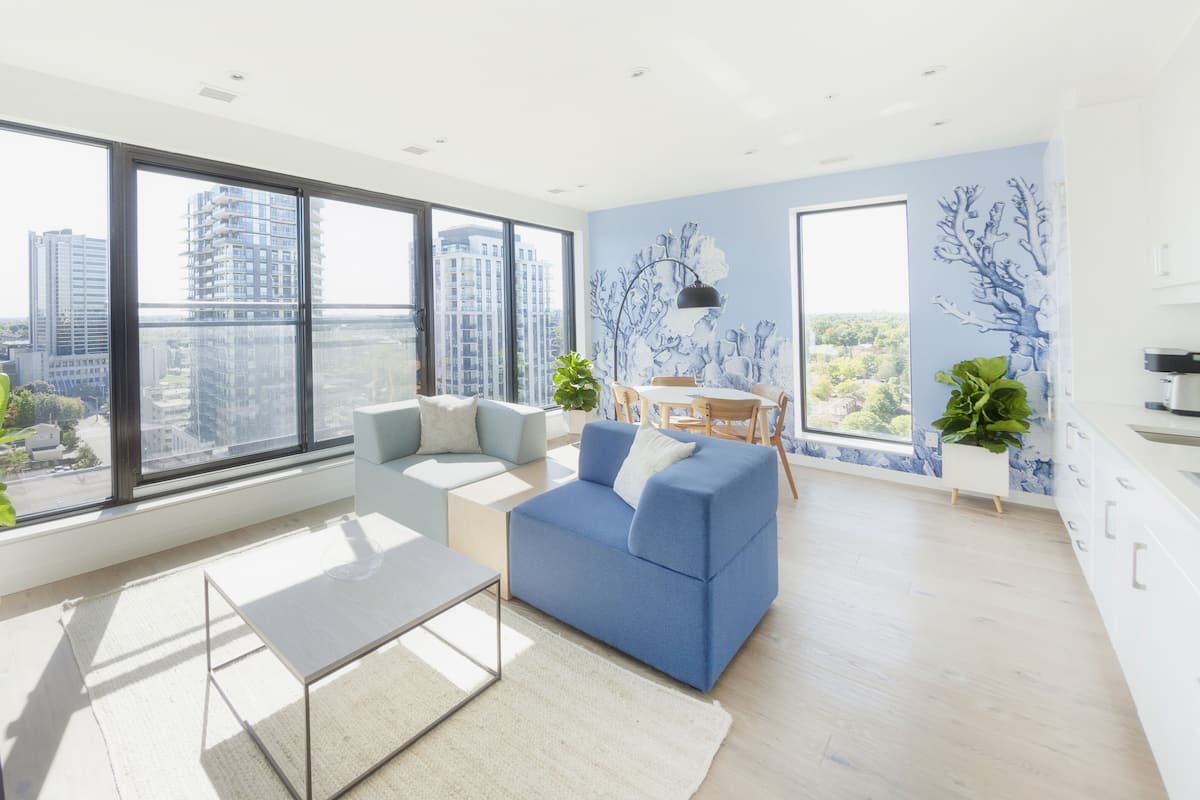 A modern living room with large windows, a blue sofa, a coffee table, and a dining area with a view of the cityscape.