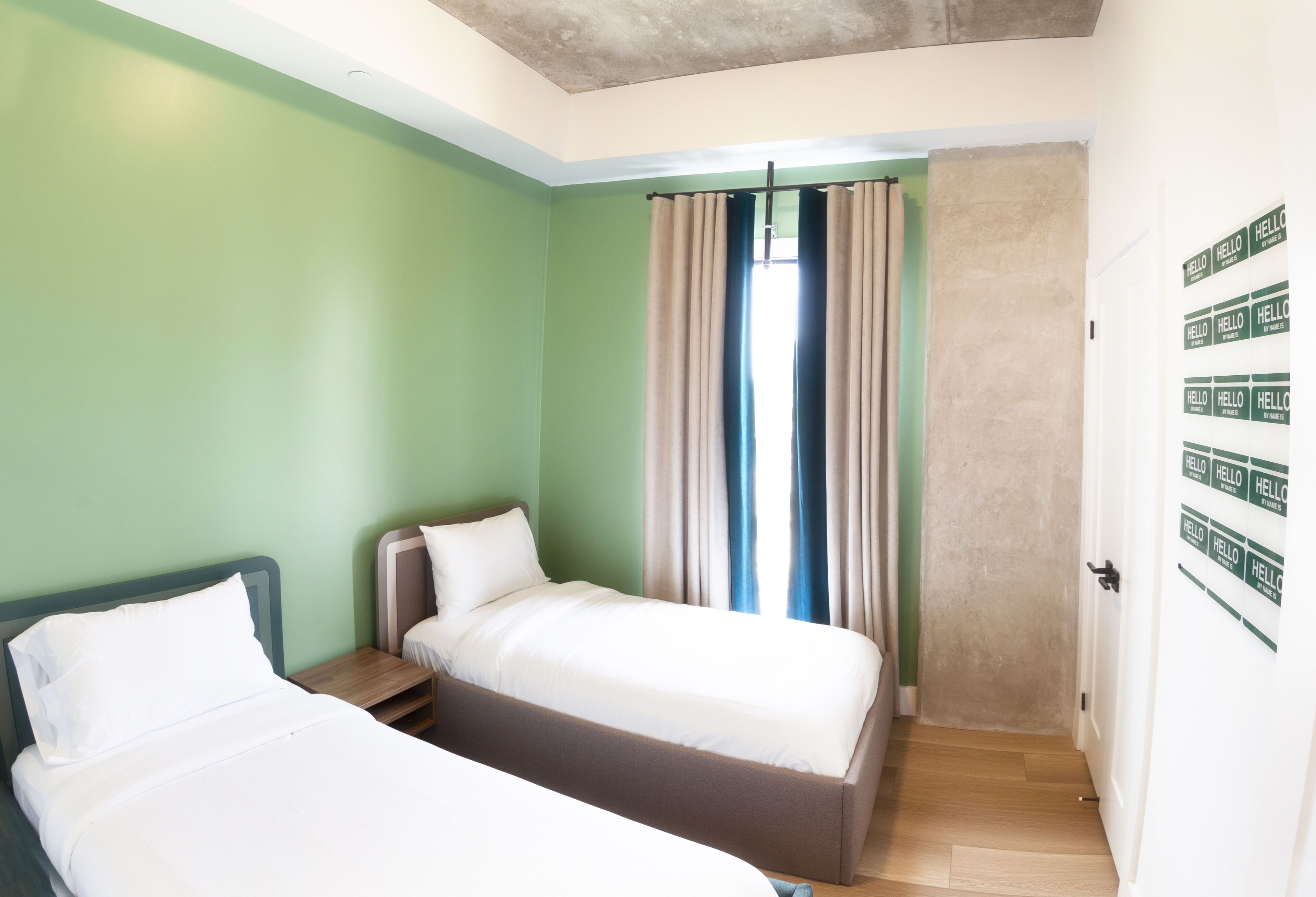 Une chambre avec deux lits, une fenêtre avec des rideaux, un plancher en bois et une porte sur laquelle est inscrit le mot « BONJOUR ».