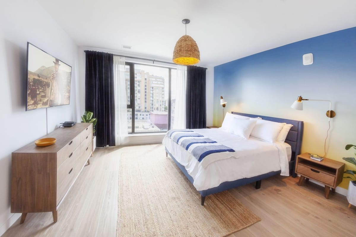 A modern hotel room with a bed, bedside tables, a TV, a large window, and a view of the city.