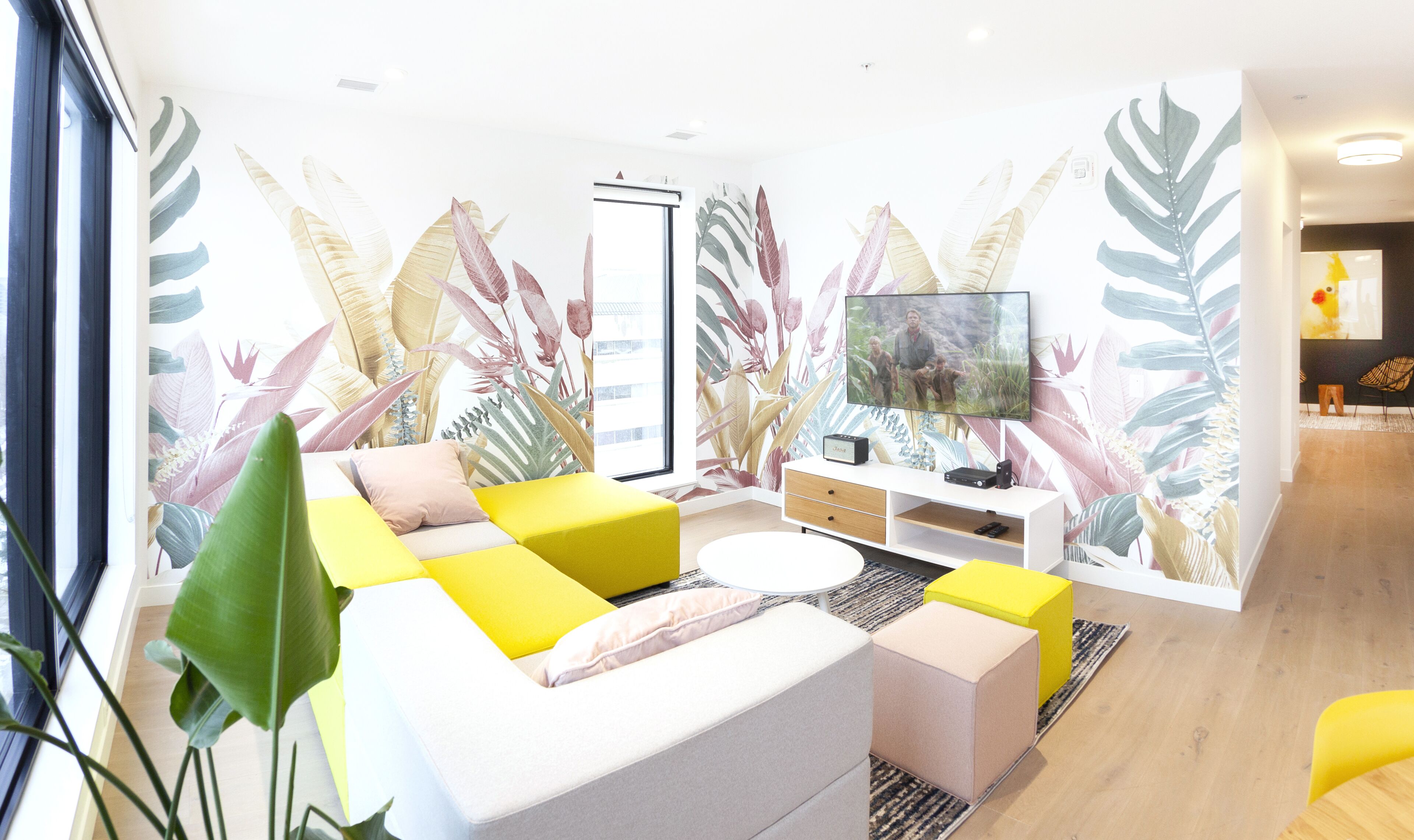 A modern living room with a sofa, a TV, and a wall with tropical leaf patterns.
