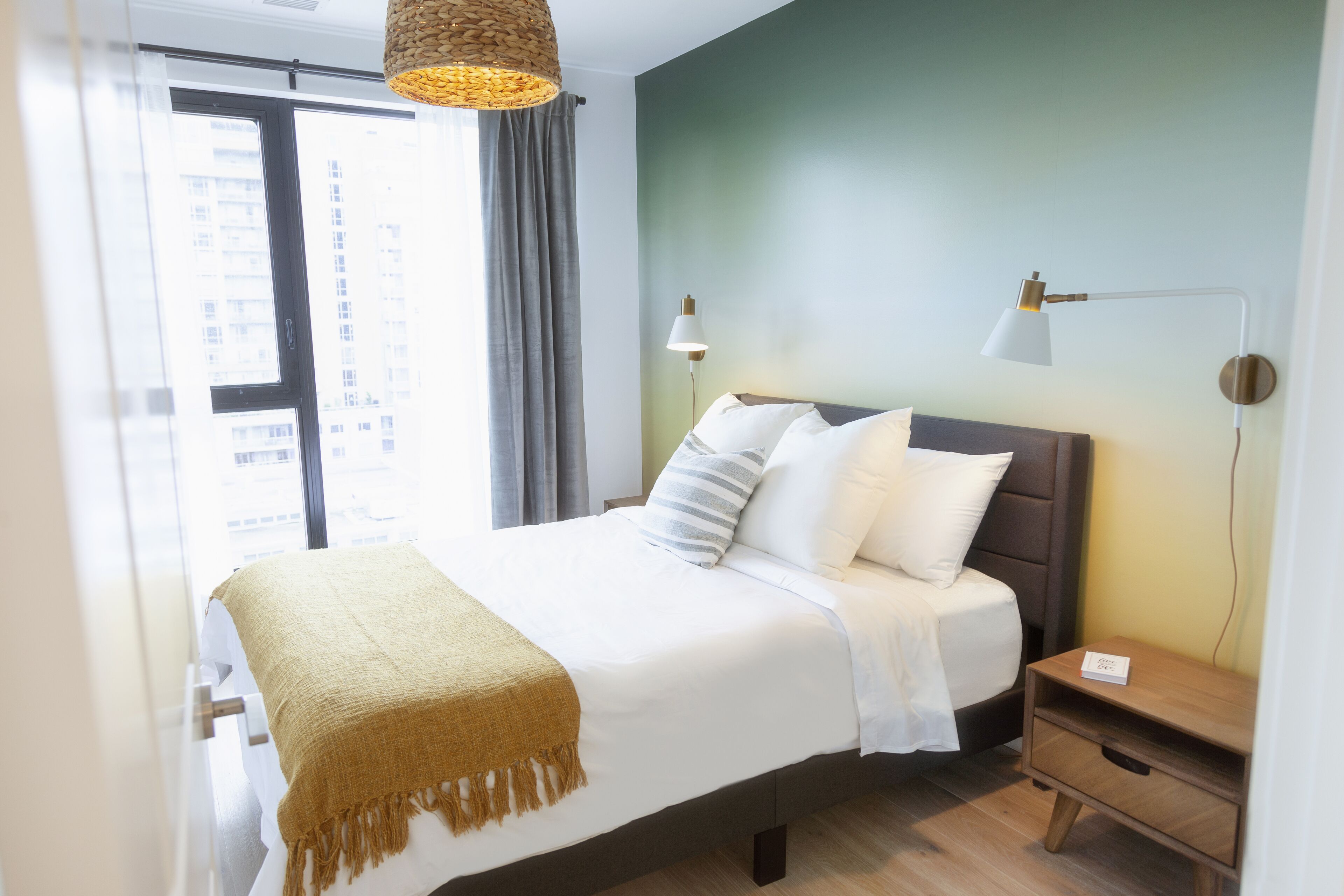 Una cama bien tendida con un edredón color beige, dos lámparas de noche y una mesita de madera pequeña con una placa.