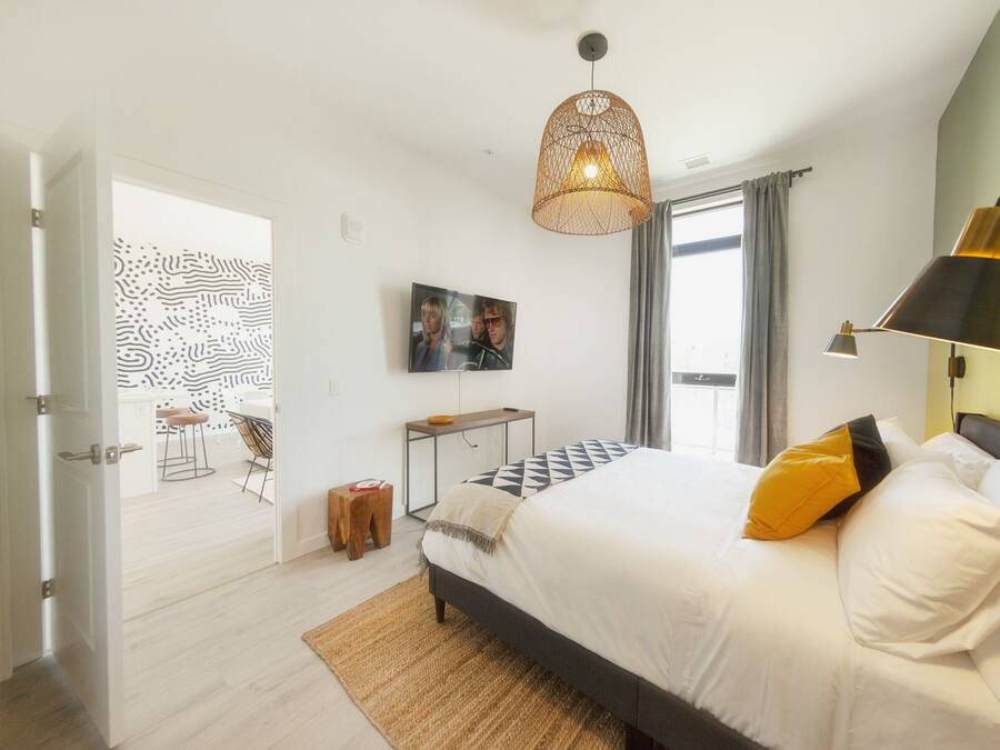 A modern bedroom with a large bed, a hanging light fixture, a bedside table, and a view of the dining area.