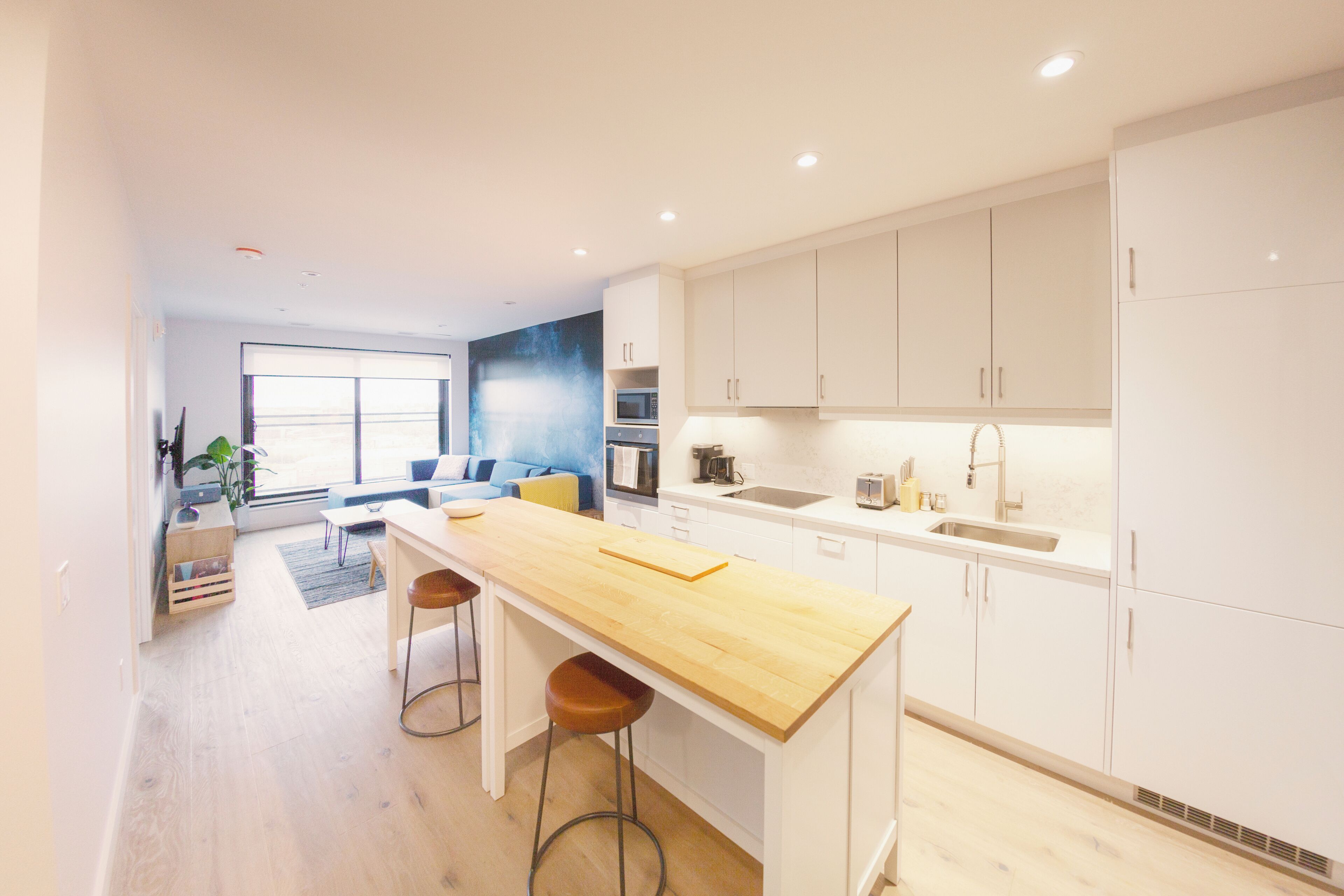 Una cocina moderna con armarios blancos, encimera de madera y horno empotrado. Hay un rincón comedor con un sofá azul y una mesita de centro.