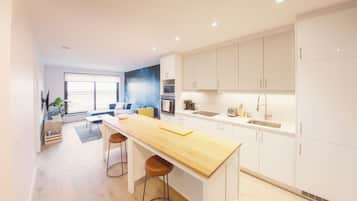 Una cocina moderna con armarios blancos, encimera de madera y horno empotrado. Hay un rincón comedor con un sofá azul y una mesita de centro.