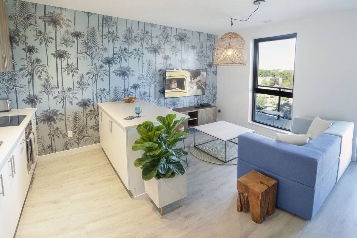 A modern kitchen with a central island, a dining area with a blue sofa, and a large window offering a view.