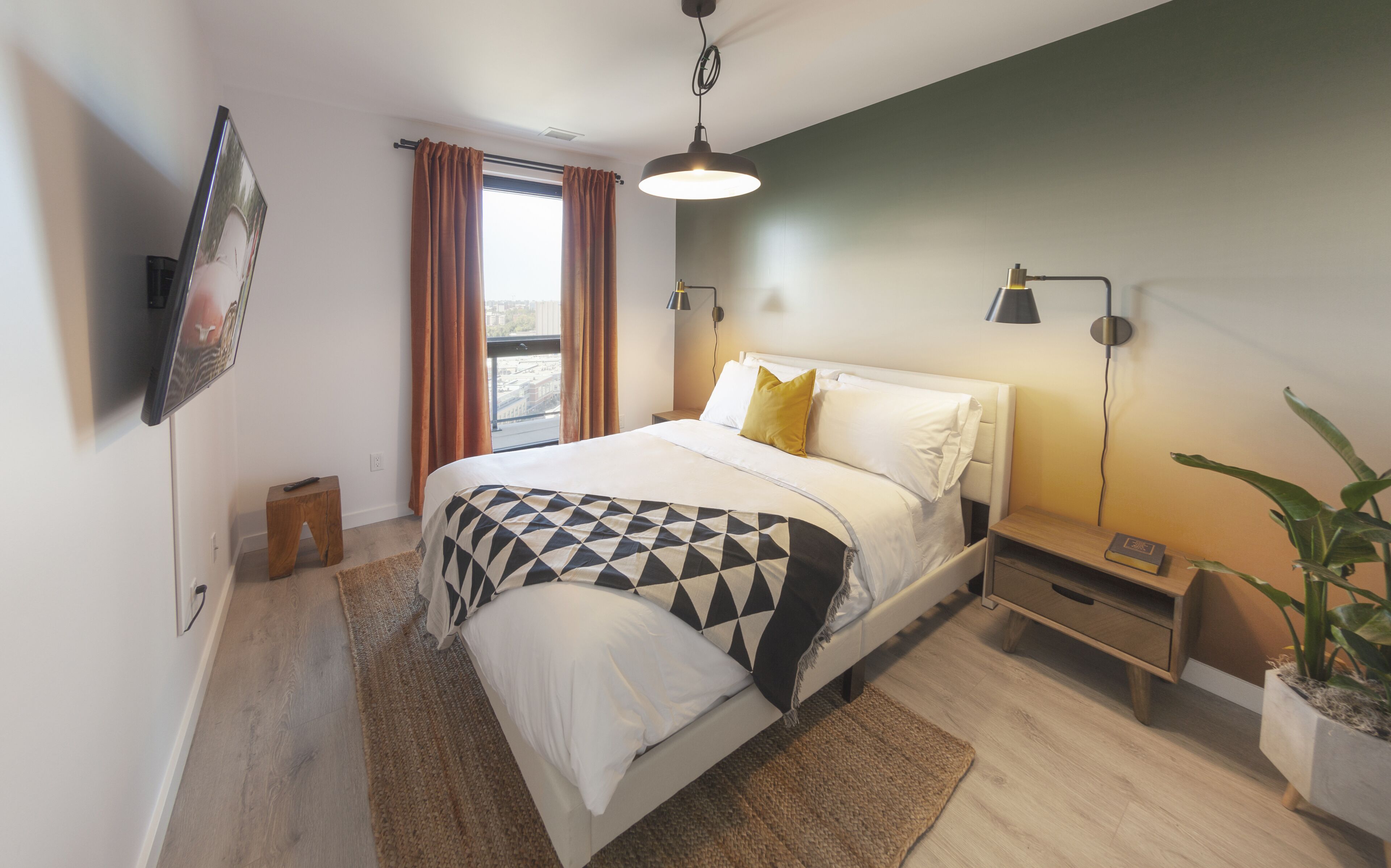 A modern bedroom with a bed, bedside lamps, a small table, a potted plant, and a window with curtains.