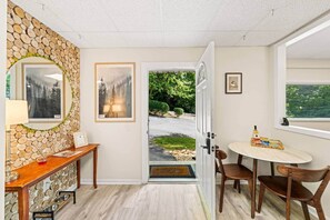Een gang met een stenen muur, een houten bank, ingelijste kunstwerken en een deur die leidt naar een tuin.