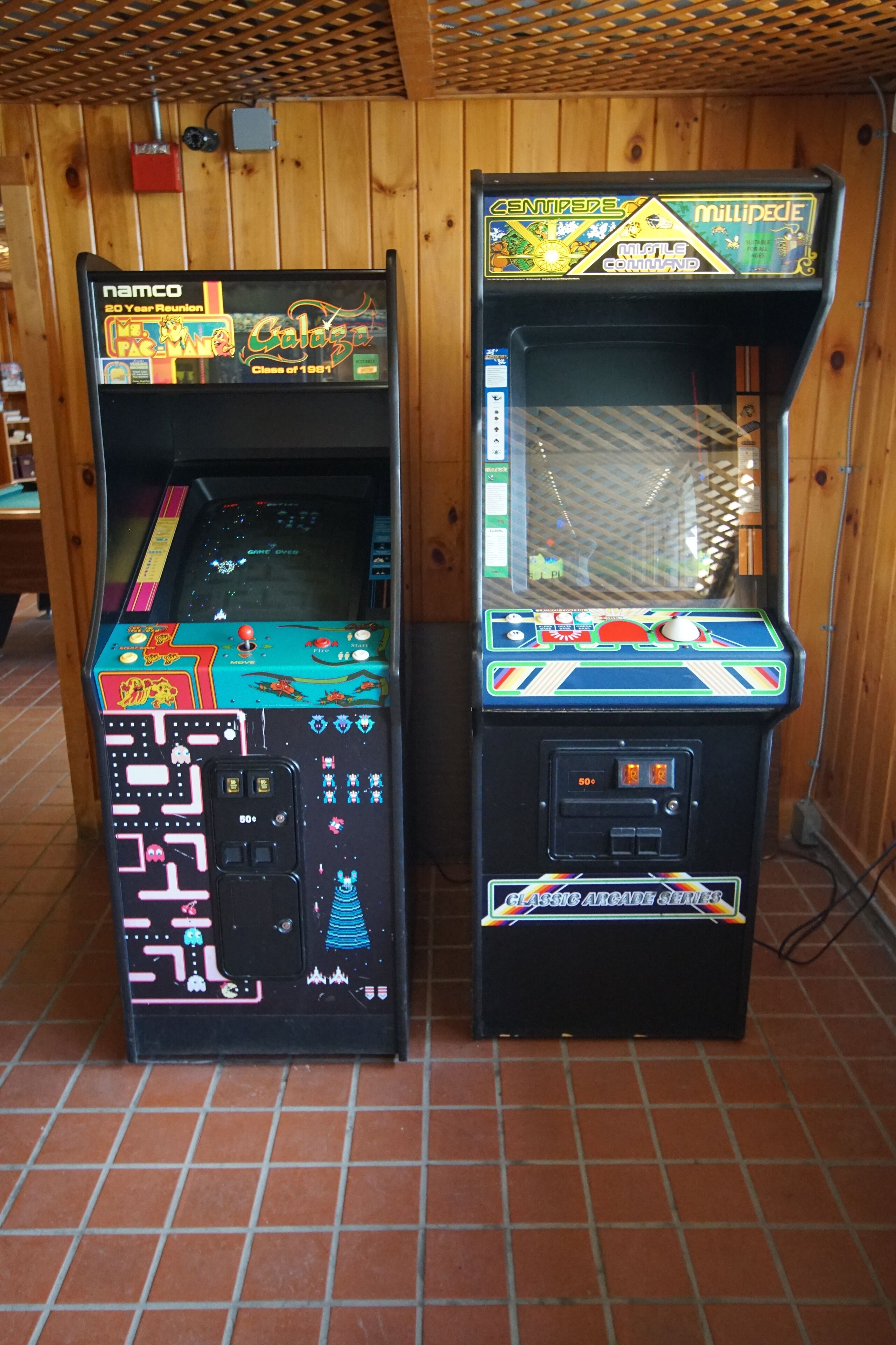 Two arcade machines, one labeled 'Namco' and the other 'Millipede', in a room with wooden walls and tiled flooring.