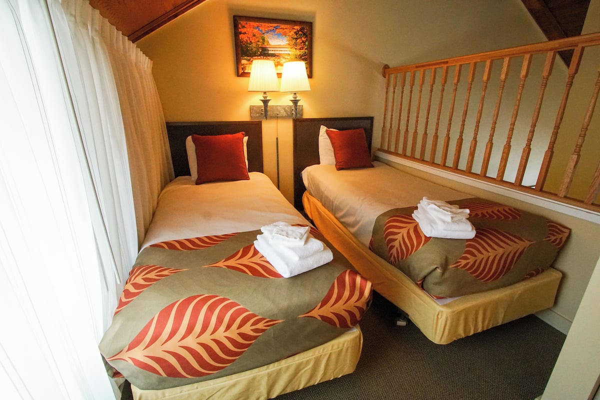 A hotel room with two single beds, each with a pillow and a folded towel. There is a painting above the beds and a wooden railing on the right.