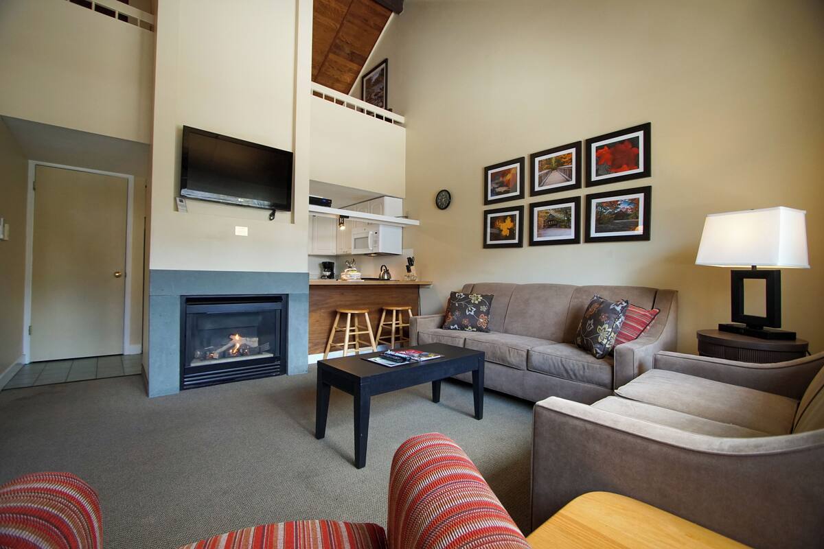 A living room with a fireplace, a sofa, a coffee table, a TV mounted on the wall, and a kitchen area in the background.