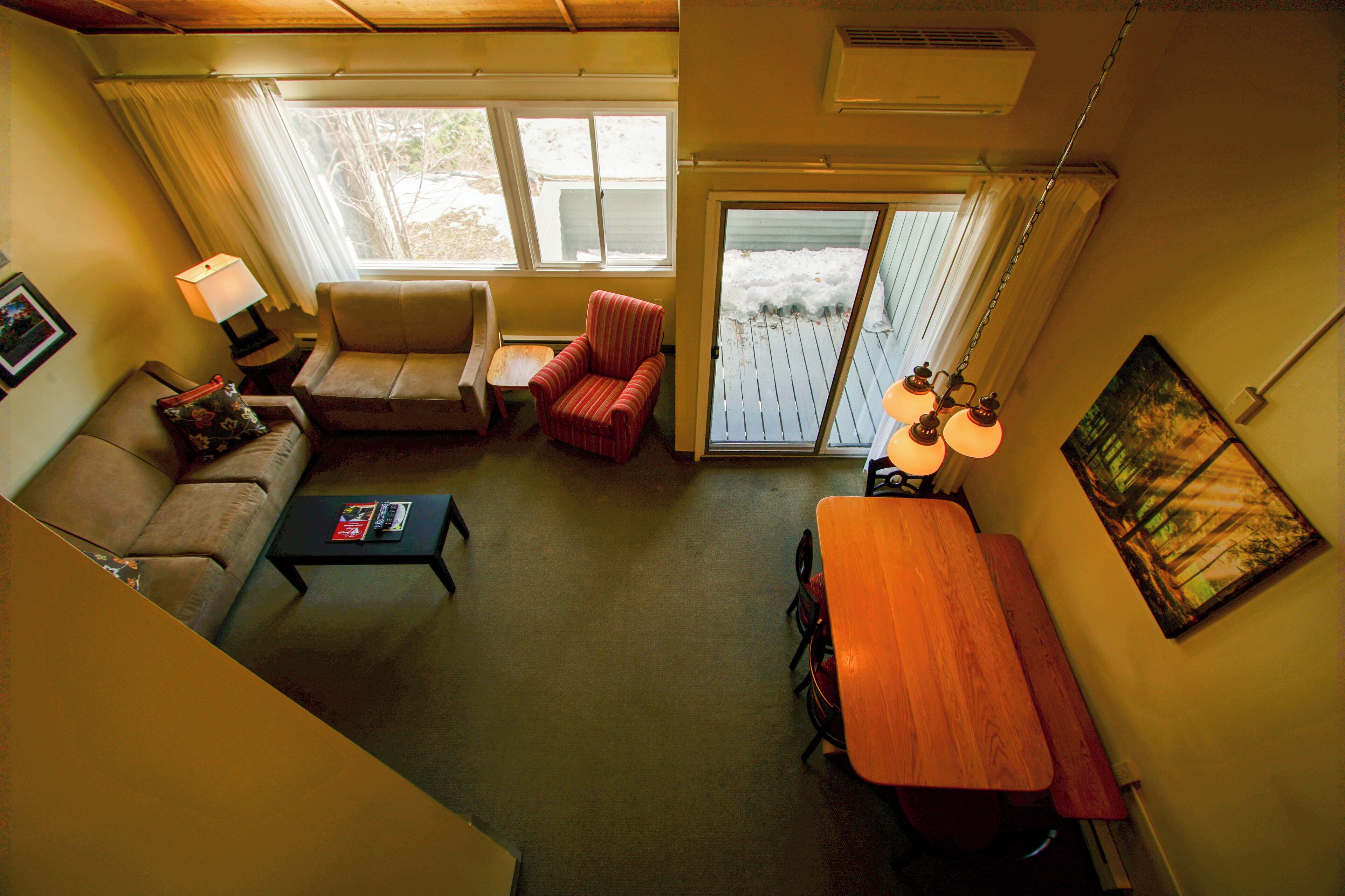 A living room with a sofa, armchair, coffee table, and wooden table. There is a window with curtains, a painting on the wall, and a ceiling fan.