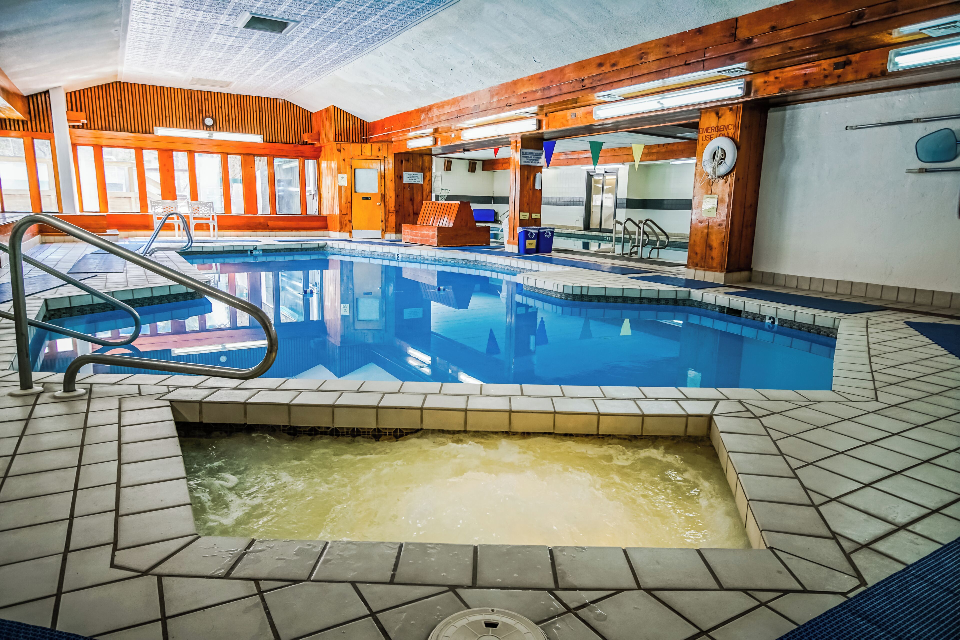 An indoor swimming pool with a tiled floor, wooden walls, and a visible emergency exit sign.