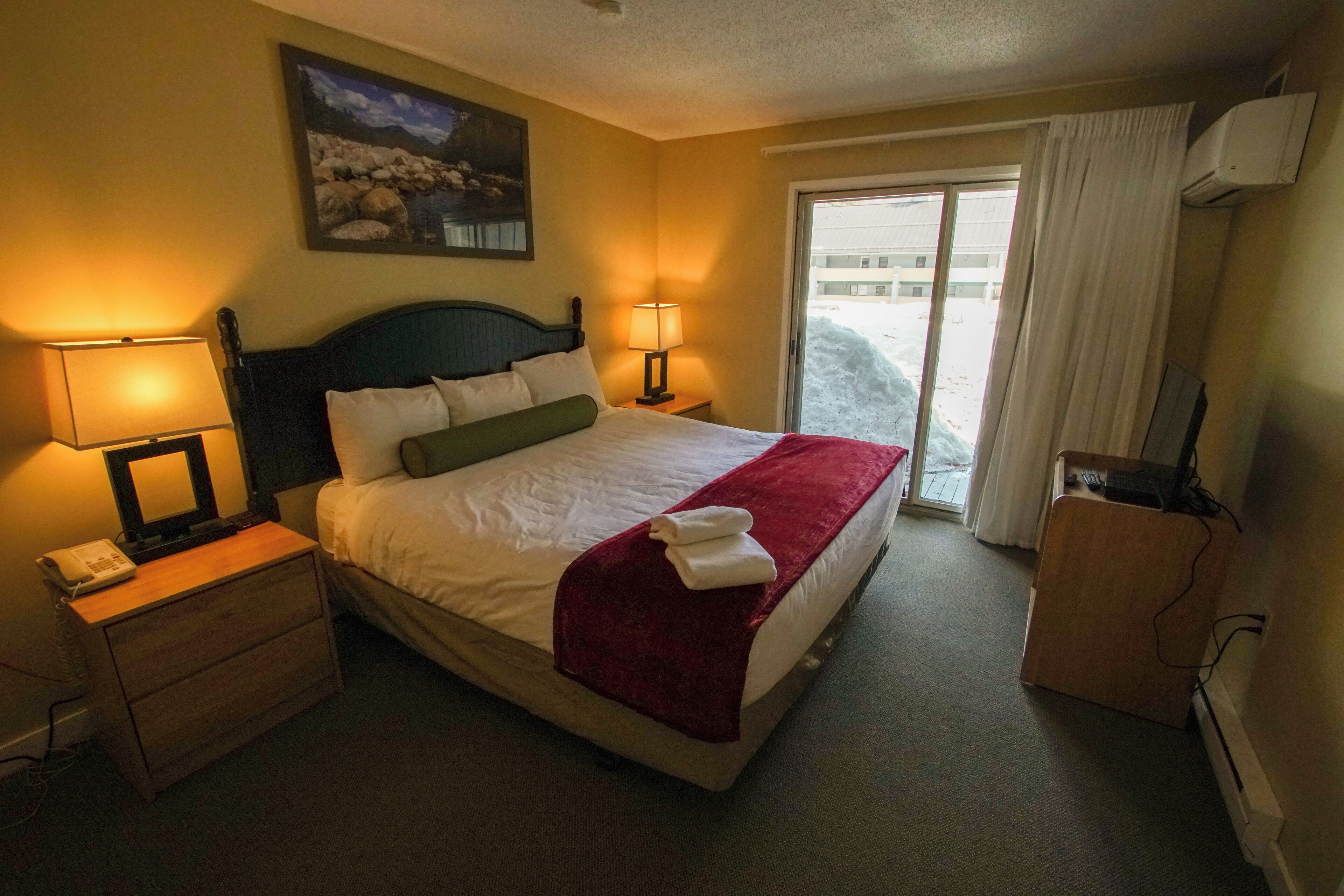 A hotel room with a bed, two bedside lamps, a telephone, a TV, and a window with curtains.