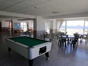 A pool table in a room with dining tables and chairs, large windows, and a view of the sea.