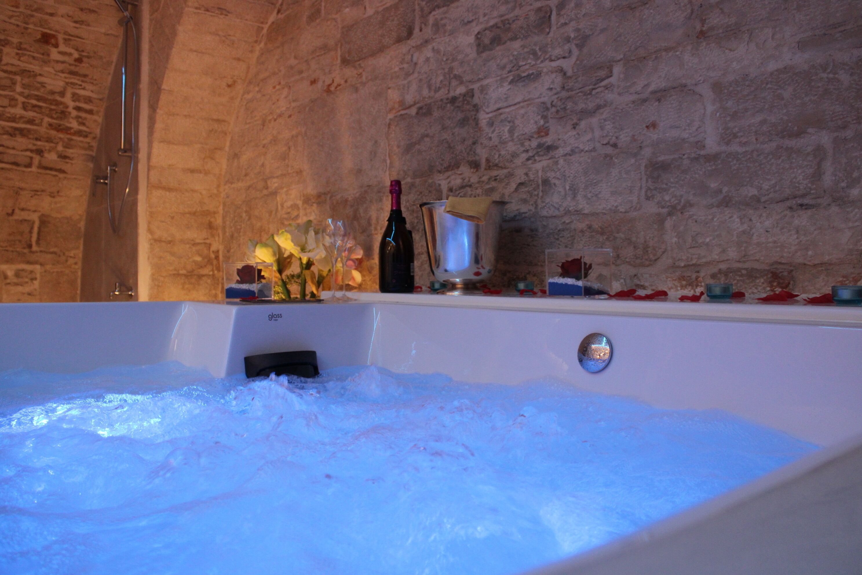 Une baignoire remplie de mousse, une bouteille de vin et un verre posé sur une étagère.