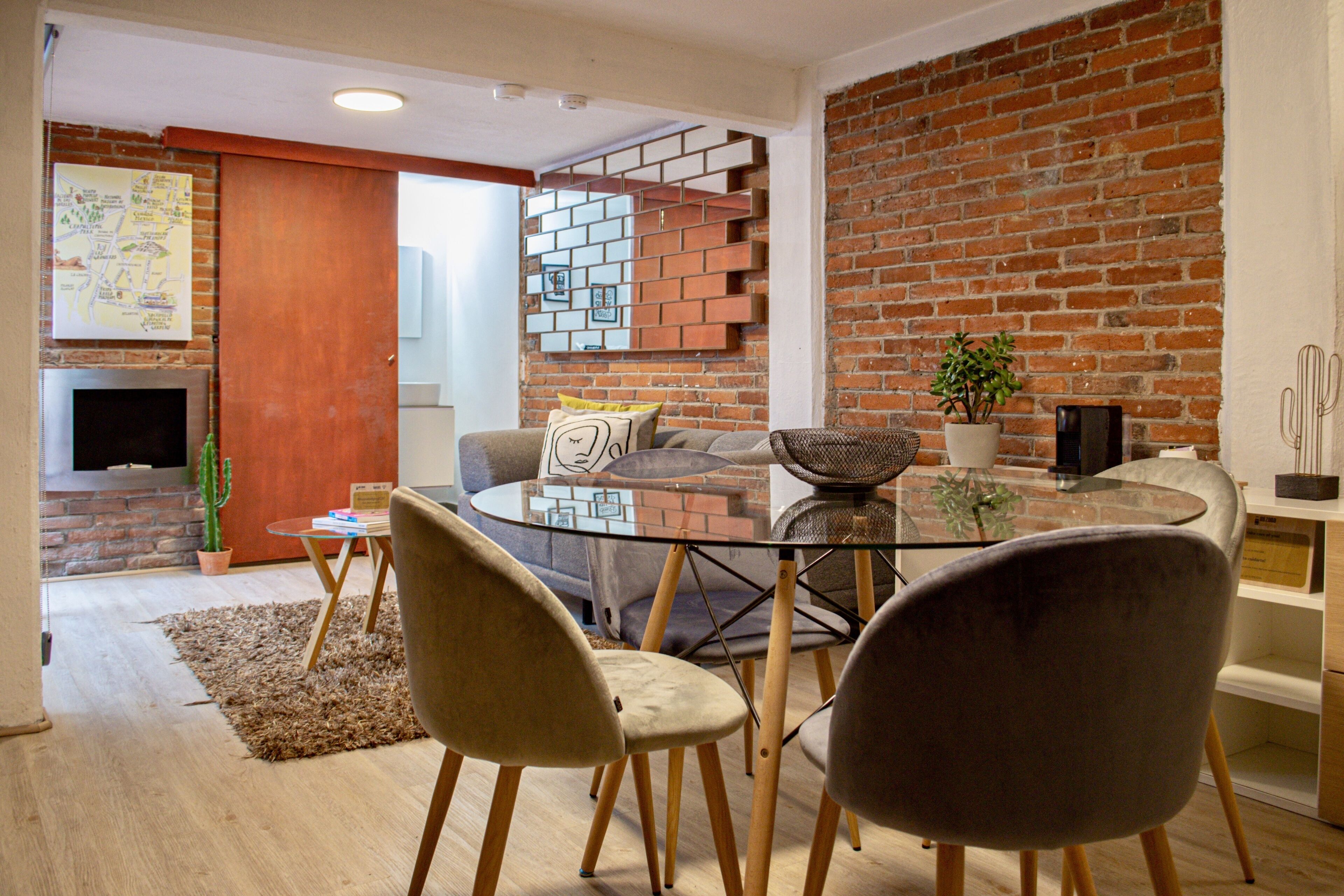 Un espacio habitable moderno con una mesa de vidrio, dos sillas, un sofá, una pared de ladrillo, un cactus y un televisor.