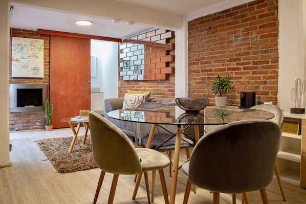 A modern living space with a glass dining table, two chairs, a sofa, a brick wall, a cactus, and a television.