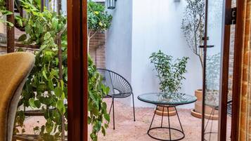 Pequeño patio con mesa de vidrio y dos sillas, rodeado de plantas en macetas y un muro de ladrillo.