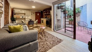 A modern living room with a grey sofa, a coffee table, and a dining area with chairs. There is a kitchen area with a stove and cabinets. The room has a large sliding glass door leading to an outdoor patio with a table and chairs. - Mi Zona Lodging Coyoacán B (Mexico City)