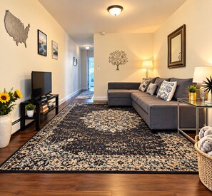 A living room with a sectional sofa, a flat-screen TV, a decorative rug, and wall art including a map and a tree. - Private One Bedroom Apartment with full kitchen, LR, full bath and private yard (Ridge)