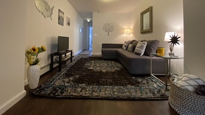 A living room with a sectional sofa, a TV, a coffee table, a rug, and a vase with flowers. - Comfy, private one Bedroom Apartment with full kitchen, living room, full bath. (Ridge)