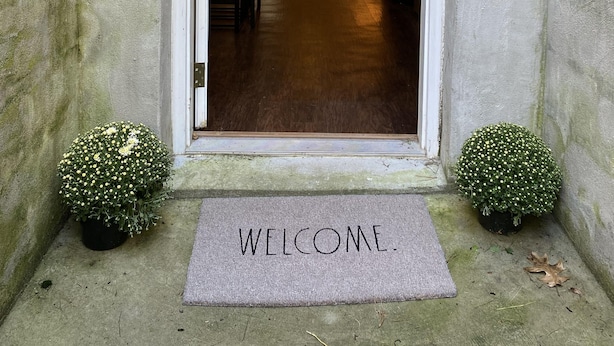 A doorway with a "WELCOME" mat, a small potted plant on each side, and a hallway leading to a living room with a couch and a lamp. - Comfy, private one Bedroom Apartment with full kitchen, living room, full bath. (Ridge)