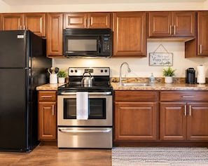A kitchen with a black refrigerator, stainless steel oven, and a microwave. There is a granite countertop, wooden cabinets, and a stainless steel sink.