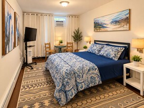 A hotel room with a bed, a desk, a chair, a television, a window with curtains, a painting on the wall, and a plant. - Private One Bedroom Apartment with full kitchen, LR, full bath and private yard (Ridge)