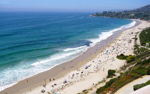 A coastal beach with a wide sandy shore, a clear blue ocean, and a hilly landscape in the background.