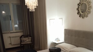 Een slaapkamer met een bed, een stoel, een nachtkastje, een spiegel en een raam met gordijnen.