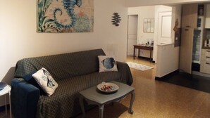 Een woonkamer met een bank, een klein tafeltje met een schaal lekkernijen, een schilderij aan de muur en een keuken op de achtergrond.