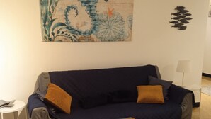 Een woonkamer met een donkerblauwe bank, twee oranje kussens, een klein rond tafeltje met een wit bord en een decoratieve wandversiering.