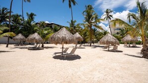 A beach with thatched umbrellas and lounge chairs, palm trees, and a building in the background. - Best View Beach front Apartment In Punta Cana (Punta Cana)