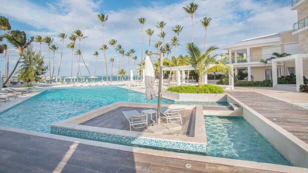A resort pool with palm trees, lounge chairs, and a clear view of the ocean. - Luxury 3BR Beachfront Condo in Punta Cana (Punta Cana)