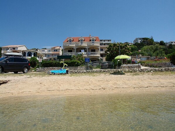 Una propiedad frente a la playa con arena, un coche aparcado y un edificio con varios balcones y techo rojo.