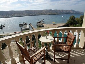 Un balcón con dos sillas de madera y una mesita pequeña, con vista a una masa de agua tranquila donde se ven barcos amarrados y una playa al fondo.