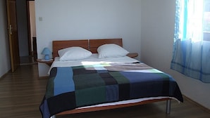 A bedroom with a wooden bed, white pillows, a blue and green quilt, a bedside table with a lamp, and a window with sheer curtains. - Pansion Gea - Superior Double Room with Balcony and Sea View (Pag)