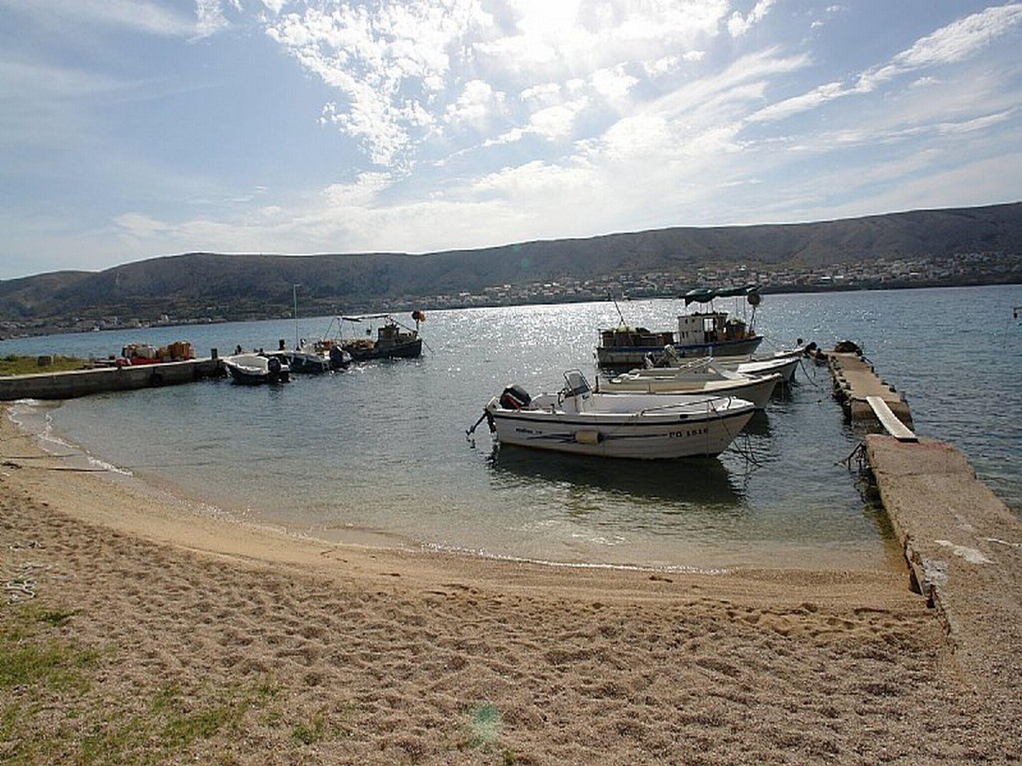 A sandy beach with boats docked at a pier, a calm body of water, and a distant shoreline with hills.