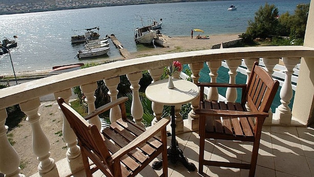 A balcony with two wooden chairs and a small table, overlooking a calm body of water with boats docked and a beach in the distance. - Pansion Gea - Superior Double Room with Balcony and Sea View (Pag)