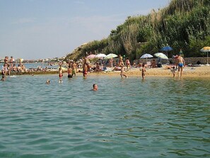 A beach with people swimming and sunbathing, umbrellas, and a stone wall in the background.