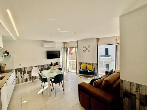 A modern living room with a sofa, a dining table with chairs, a TV mounted on the wall, and a large window.