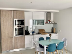 A modern kitchen with white cabinets, a Bosch oven, a microwave, a glass dining table with teal chairs, and a small shelf with decorative items.