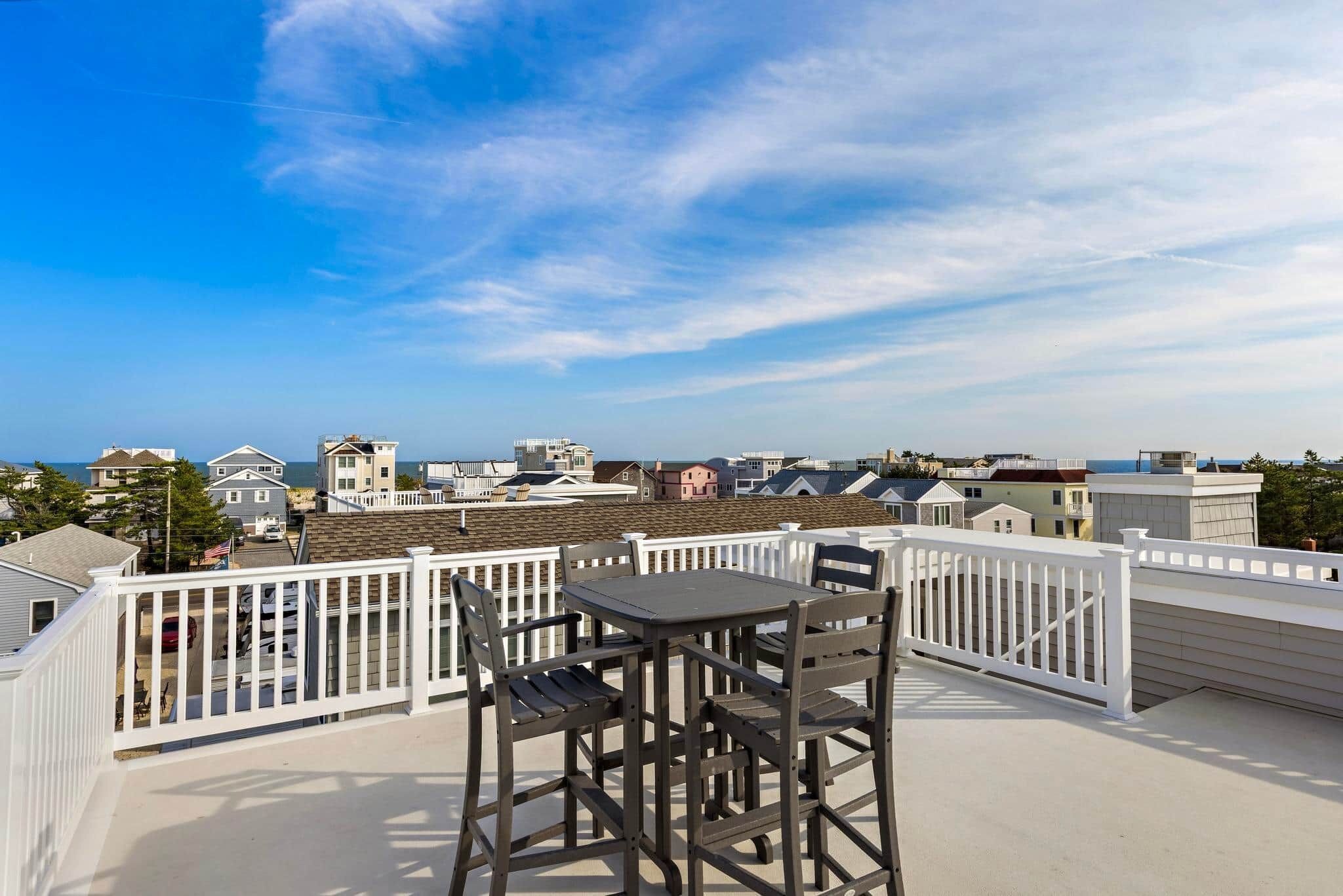 Terraço no telhado com mesa e cadeiras, guarda-corpo branco e vista para o oceano e edifícios próximos.