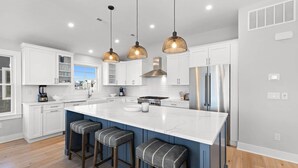 A modern kitchen with white cabinets, a large island with bar stools, stainless steel appliances, and pendant lighting. - Ultimate Beach Getaway (Long Beach)