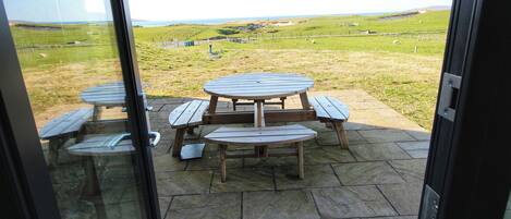 A patio with a round table and benches, overlooking a grassy field with a fence and distant hills.